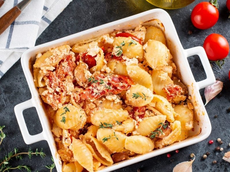 Baked Feta Pasta in einer weißten Auflaufform mit frischem Thymian auf einem graunen Schiefertisch mit frischem Pfeffer, Knoblauch und Tomaten als Deko.