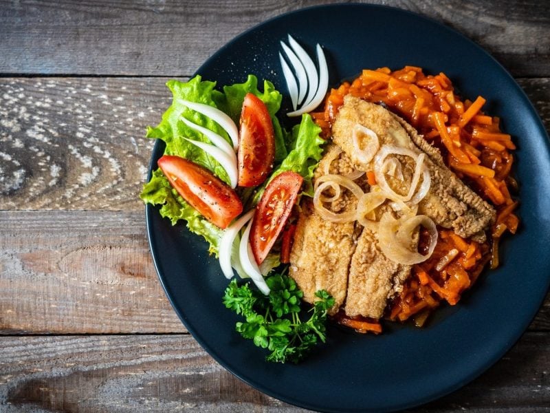 Gebratenes Fischfilet mit Tomatensauce : ein Teller mit Tomaten-Möhren-Soße, Fisch und Salat auf einem Holztisch.