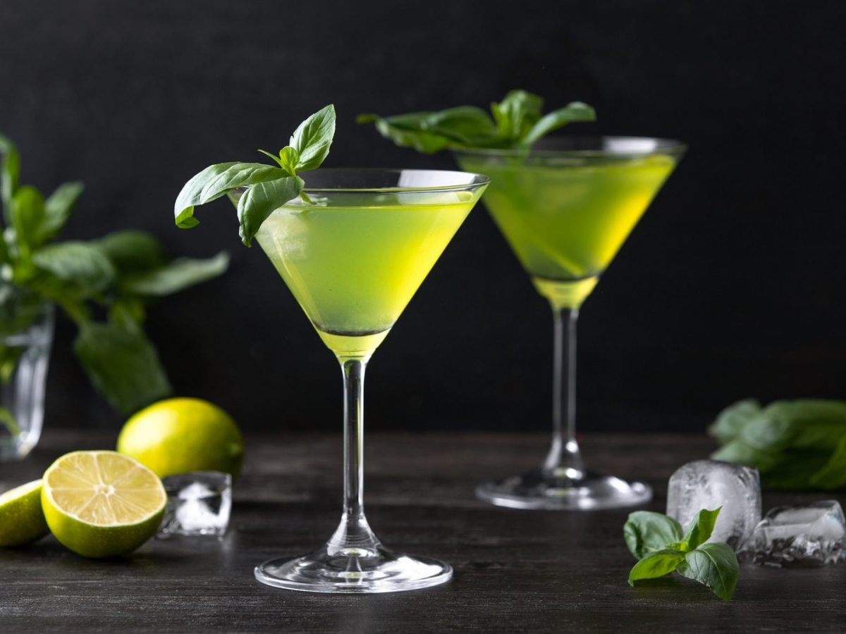 Zwei Gläser mit Gin-Basil-Smash stehen auf einem dunklen Untergrund vor einer schwarzen Wand. Um die Gläser herum liegen Basilikumblätter, angeschnittene Zitronen und einige Eiswürfel. Die Cocktails sind mit Basilikumblättern angerichtet.