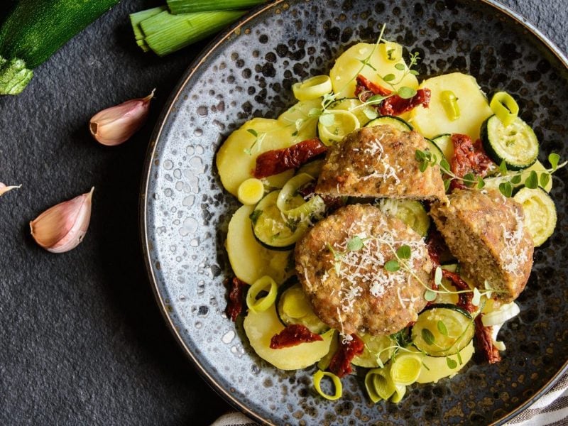 Griechische Hackbällchen auf einem Teller mit Zucchini und Kartoffelscheiben, dazu getrocknene Tomaten, Lauch und Kresse. Daneben Lauchstangen, eine Zucchini und Knoblauchzehen.