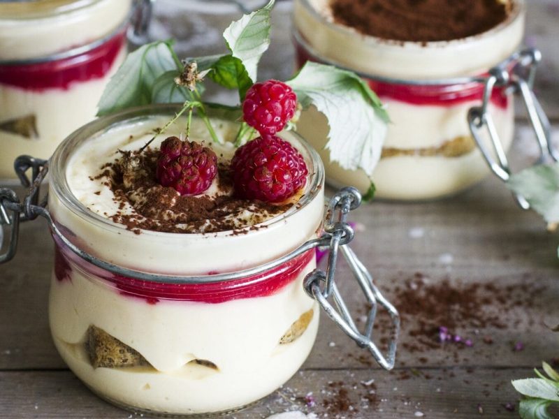 Mehrere verschließbare Gläser stehen auf einem grauen Tisch. Die Gläser sind gefüllt mit Himbeer-Tiramisu und bestreut mit Kakaopulver. Neben den Gläsern ist etwas Kakaopulver auf dem Tisch verstreut. Einige Himbeeren mit Blättern liegen dekorativ am Bildrand.