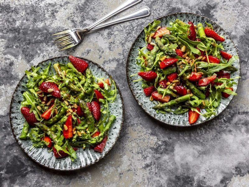 Bunter Spargelsalat mit Erdbeeren und Pinienkernen auf zwei Tellern auf einem grauen Steintisch neben zwei Gabeln.