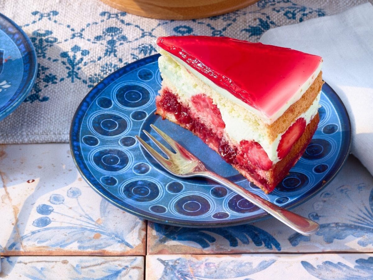 Sahnetorte mit Erdbeerbowle-Füllung mit frischen Erdbeeren und einem Erdbeerbowlen-Guss auf einem verzierten blauen Teller mit einer Gabel auf einem blauen Fliesentisch neben Papierservietten.