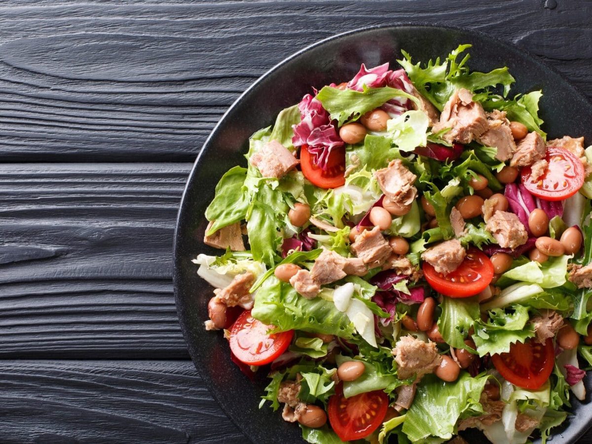 Eine Schüssel mit Thunfisch Tomaten Salat steht auf einem schwarzen Teller auf einem schwarzen Holztisch. Im Salat sind einige Bohnen verteilt.