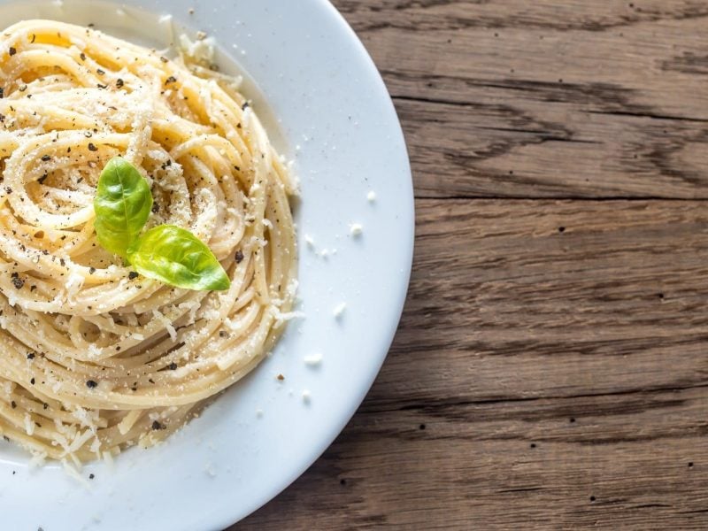 Pasta Cacio e Pepe in einem weißen Teller mit frischem Basilikum, geriebenem Parmesan und gemahlenem Pfeffer auf einem hellen Holztisch.