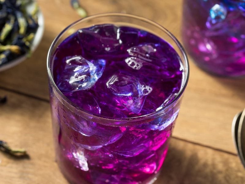 Ein Glas mit Schmetterlingsblüten Limonade mit Eiswürfeln auf einem hellen Holztisch mit frischen Schmetterlingsblüten und zwei Löffeln.