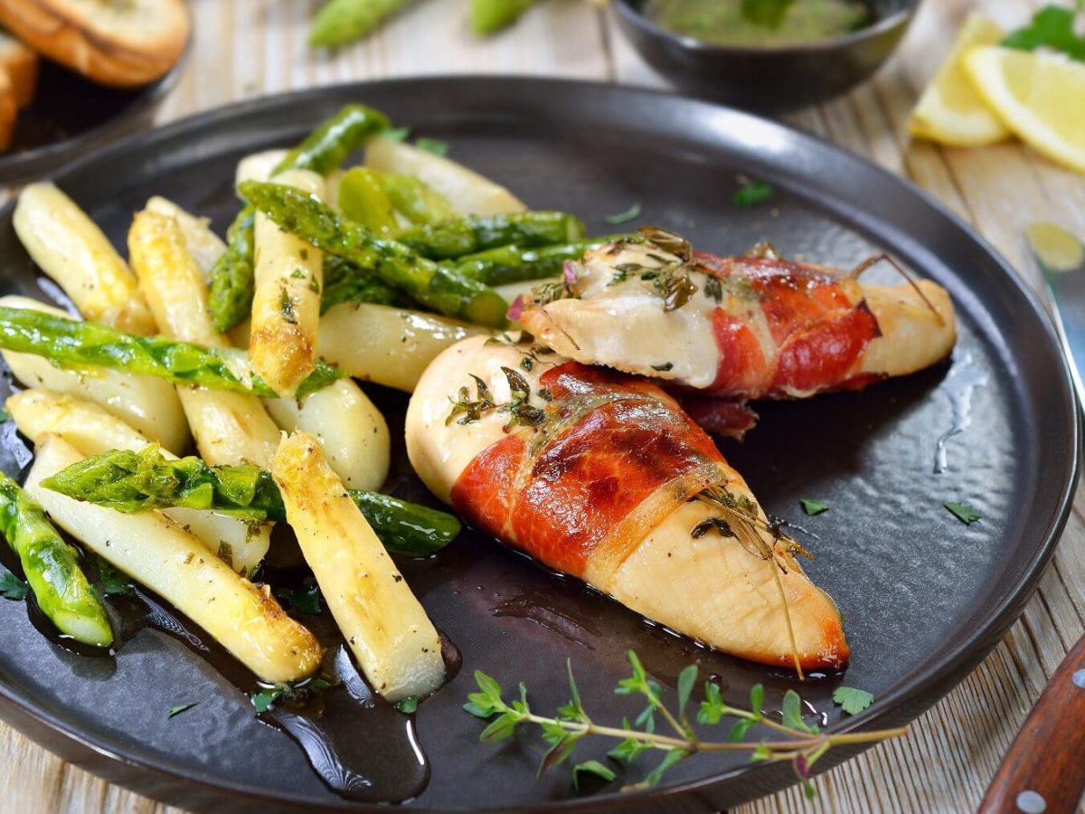 Gebratener grüner und weißer Spargel mit in Schinken gewickelter Hähnchenbrust auf einem schwarzen Teller neben Besteckt auf einem hellen Holztisch.