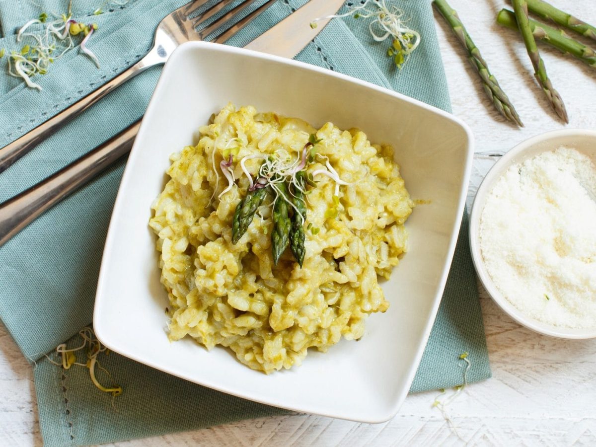 Risotto mit grünem Spargel: ein Teller mit Risotto und grünem Spargel auf einer Serviette, daneben eine Schüssel Parmesan, Besteck und ein paar Spargelstangen.