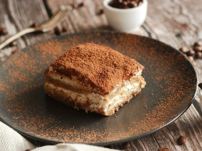 Ein Stück Tiramisu mit Baileys auf einem schwarzen Teller, mit Kakao-Pulver bestäubt, neben Schüsseln voller Kaffeebohnen