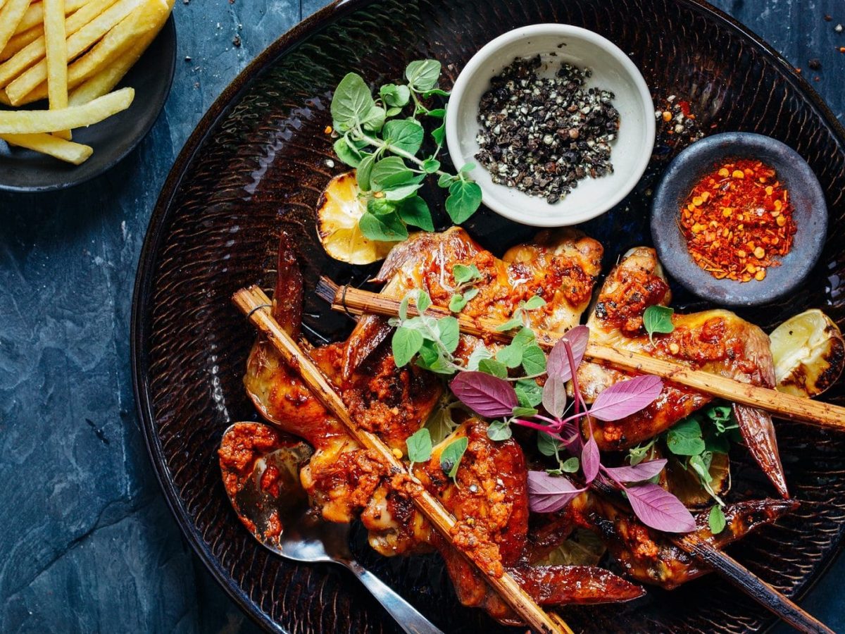 Thailändisches Hähnchen mit Teriyakisoße: ein Teller mit Hähnchenspießen, Gemüse und einer Schale mit Pfeffer- und Chilikörner. Daneben eine Schüssel Pommes.