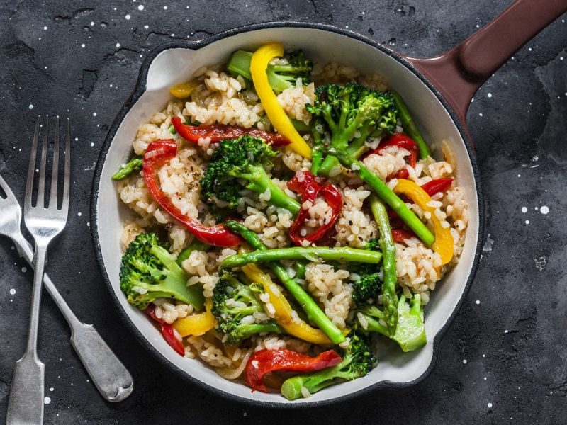 Vegetarische Reispfanne mit Paprika und Brokkoli, daneben zwei Gabeln auf dunklem Untergrund.