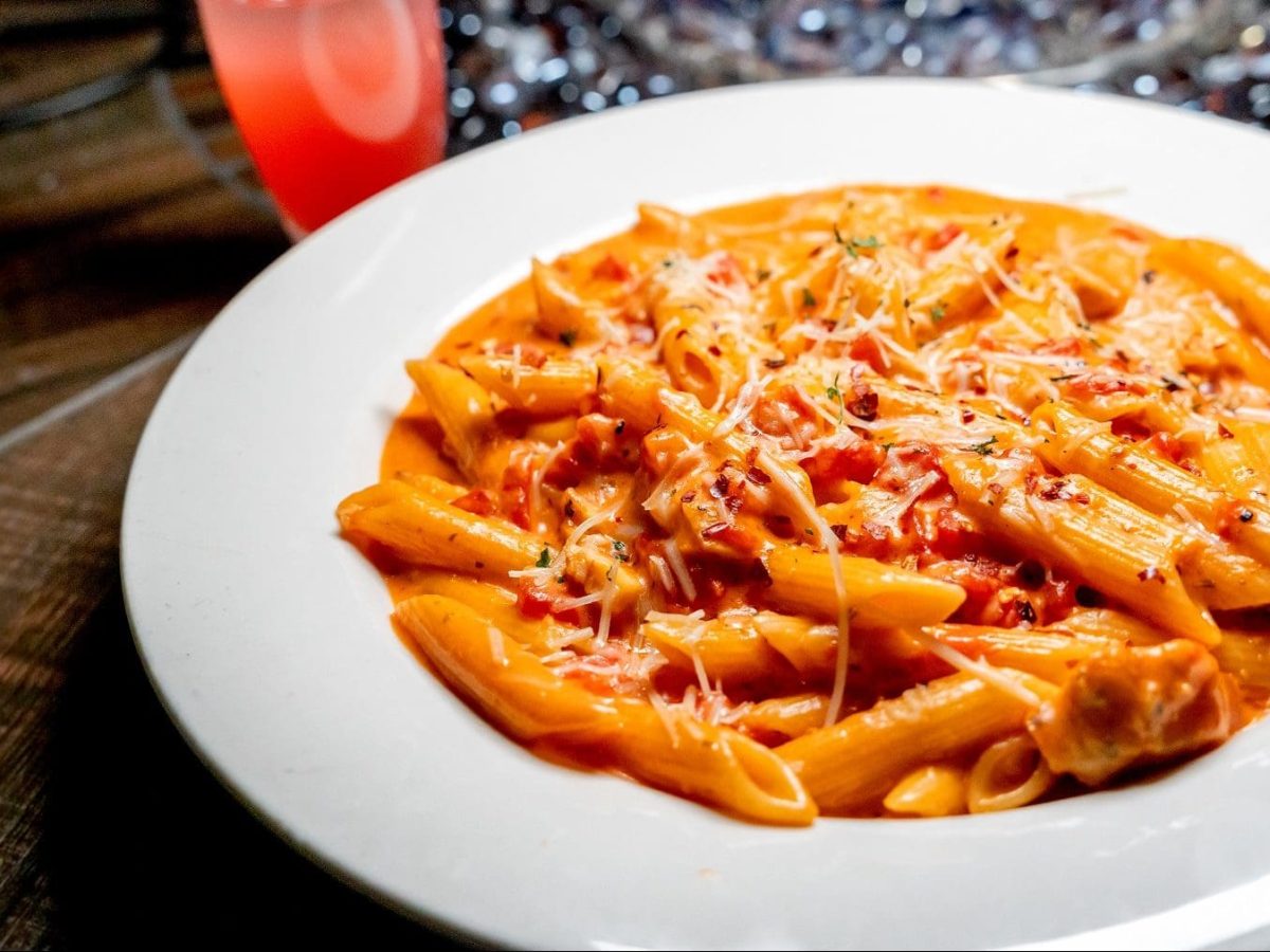 Italianische Pasta alla Vodka in einem weißen Teller auf einem dunklen Holztisch neben einem Cocktail und einer brennenden Feuerschale.