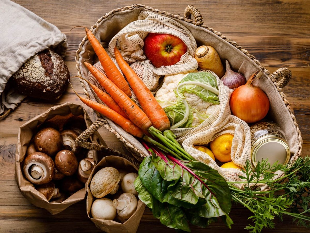 Eine große Tasche, zwei Papiertüten und ein Beutel stehen auf einem dunkelbraunen Holztisch. Die Tasche ist gefüllt mit einer Variation aus Obst und Gemüse, darunter unter Anderem Karotten, Blumenkohl und Apfel. Die Papiertüten sind gefüllt mit Pilzen. In dem Beutel am linken Bildrand befindet sich ein Brot.