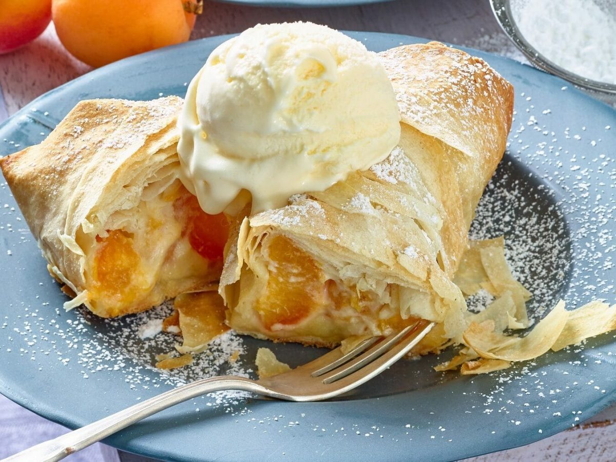 Eine angeschnittene Mini-Aprikosen-Strudelrolle liegt auf einem blauen Teller auf einem hölzernen Untergrund. Neben der Rolle liegt eine silberne Gabel. Die Strudelrolle ist garniert mit Puderzucker und einer Kugel Vanilleeis. Im Hintergrund liegt ein Sieb mit Puderzucker und zwei Aprikosen.