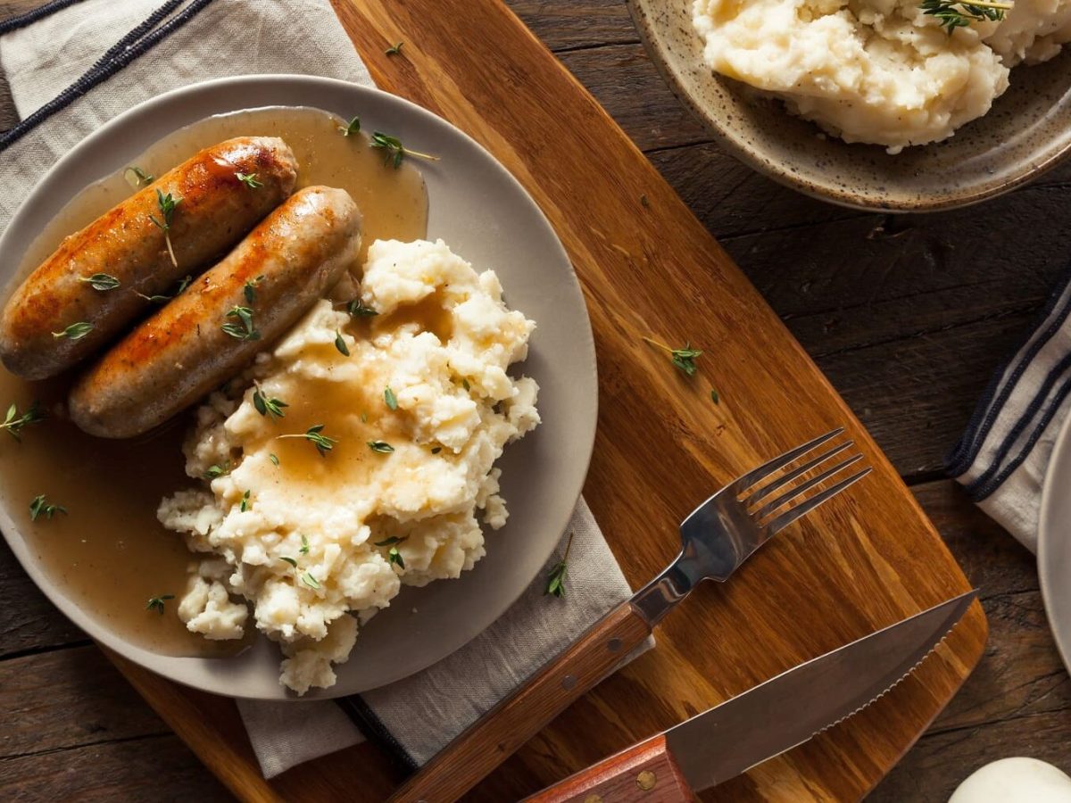 Bangers and Mash von oben auf einem hellrauen Tellern neben Besteck auf einem dunklen Holztisch.