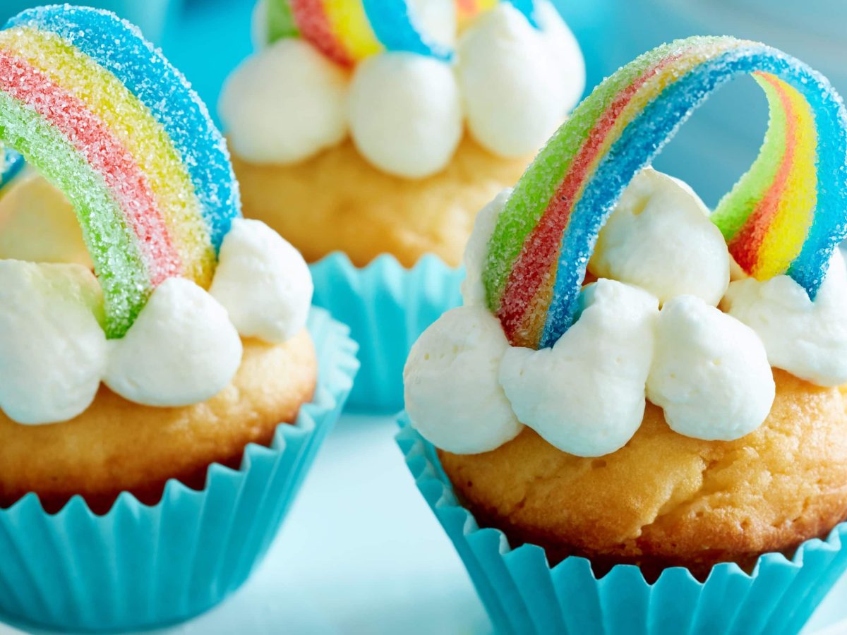 Rainbow-cupcakes in blauen Förmchen mit Regenbogen und Sahne auf einem weißen Ständer.