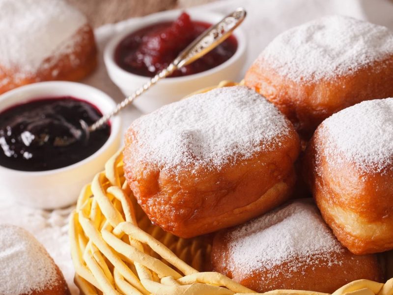 Gebackene Beignets mit Puderzucker in einem hellen Korb vor Schüsseln mit Marmelade auf einem Tisch mit weißer Tischdecke.