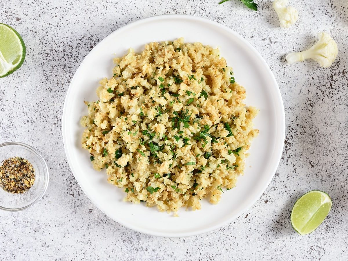 Ein Teller mit gebratenem und mit Kräutern gewürztem Blumenkohlreis neben Limetten Scheiben und Blumenkohlröschen, von oben vor einem grauen Hintergrund.