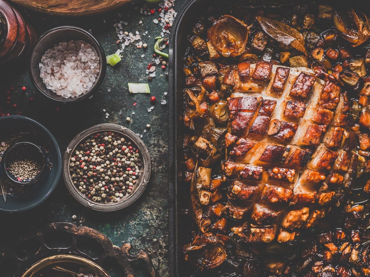 Ein Braten mit ziselierter Kruste liegt auf einer Schieferplatte mit allerlei Gewürzen drumherum