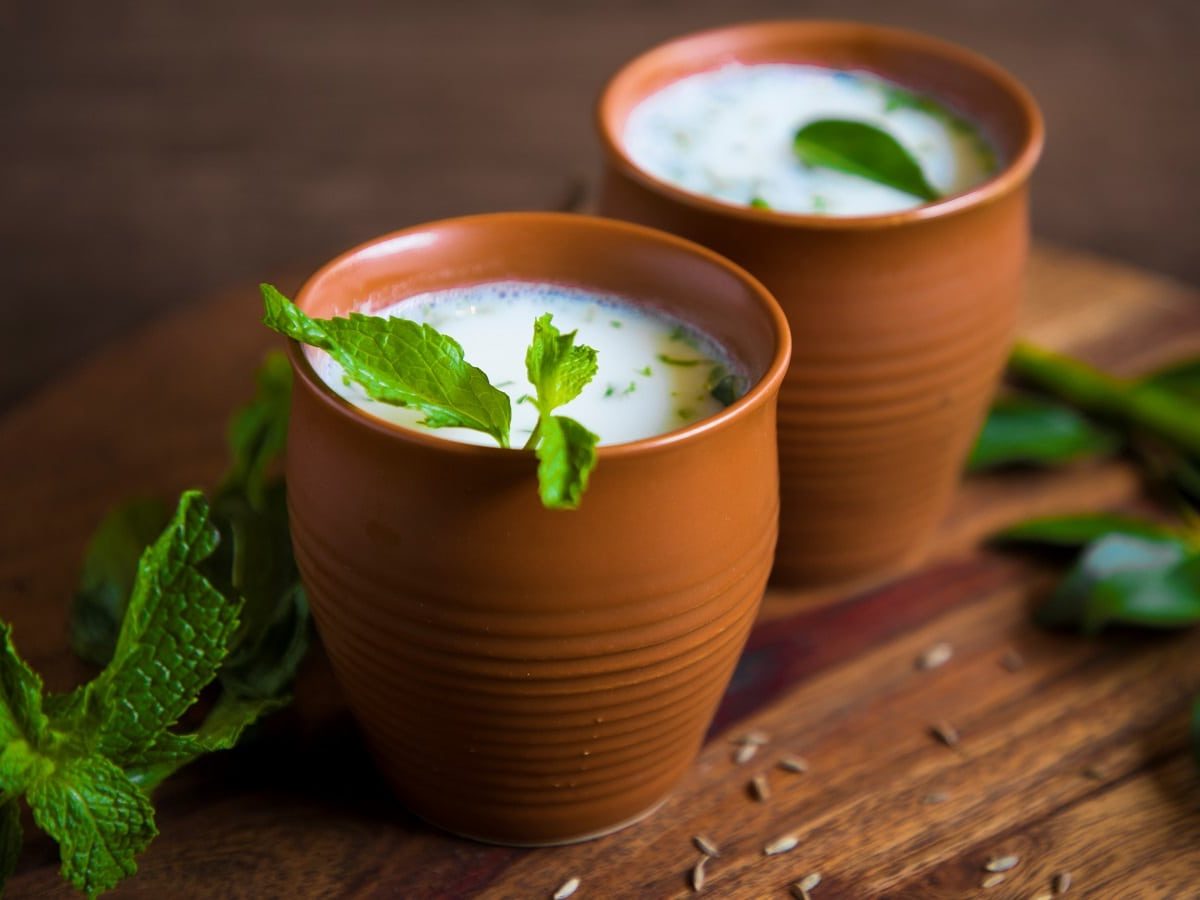 Zwei Ton-Tassen gefüllt mit dem schaumigen Buttermilch-Lassi mit Minze garniert, auf einem Holzbrett von der Seite.