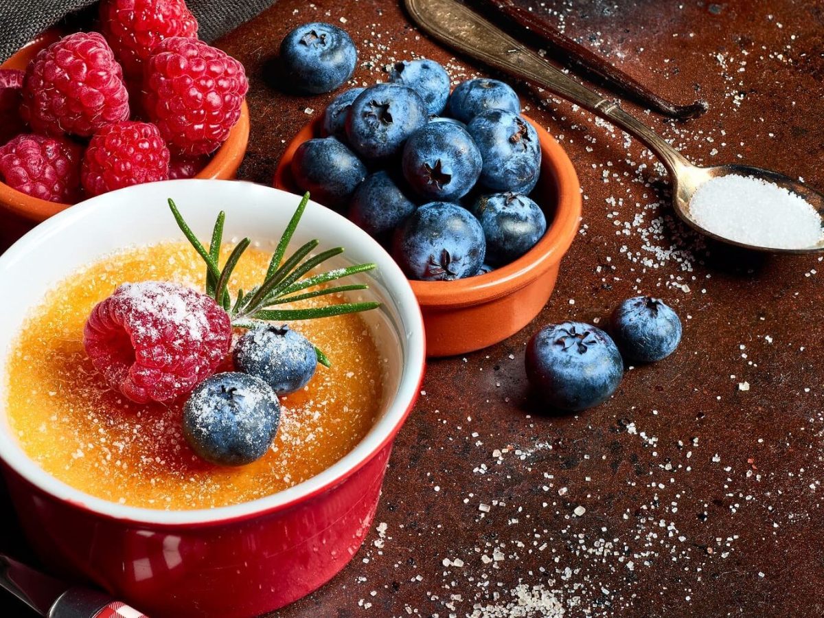 Crème Brûlée mit Kardamom und Banane in einer roten Form mit frischen Früchten auf einem dunklen Tisch der mit Zucker bestreut ist.