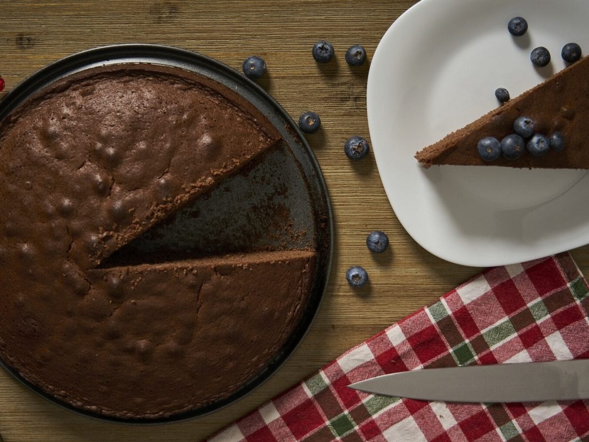 Ein angeschnittener Depression-Cake auf einem dunklen Kuchenteller steht auf einem braunen Tisch. Rechts neben dem Kuchen steht ein weißer Teller mit einem Stück des Kuchens. unter dem Teller liegt ein kariertes Handtuch mit einem Kuchenmesser.