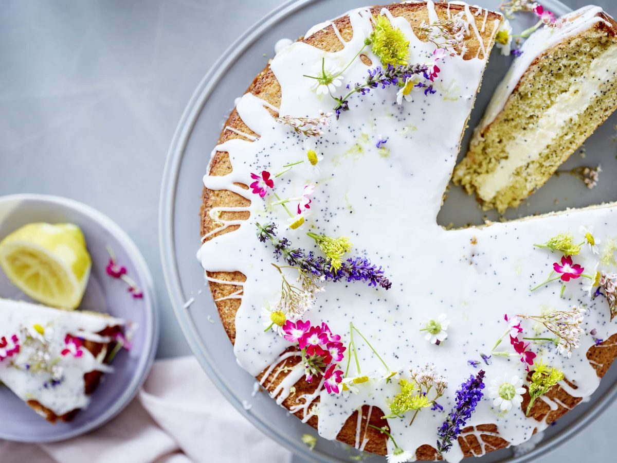 Drizzle-Kuchen mit Zitrone und Mohn verziert mit Blumen. Das Ganze auf einem Tortenhalter und daneben ein Stück auf einem Teller mit einer Zitrone.