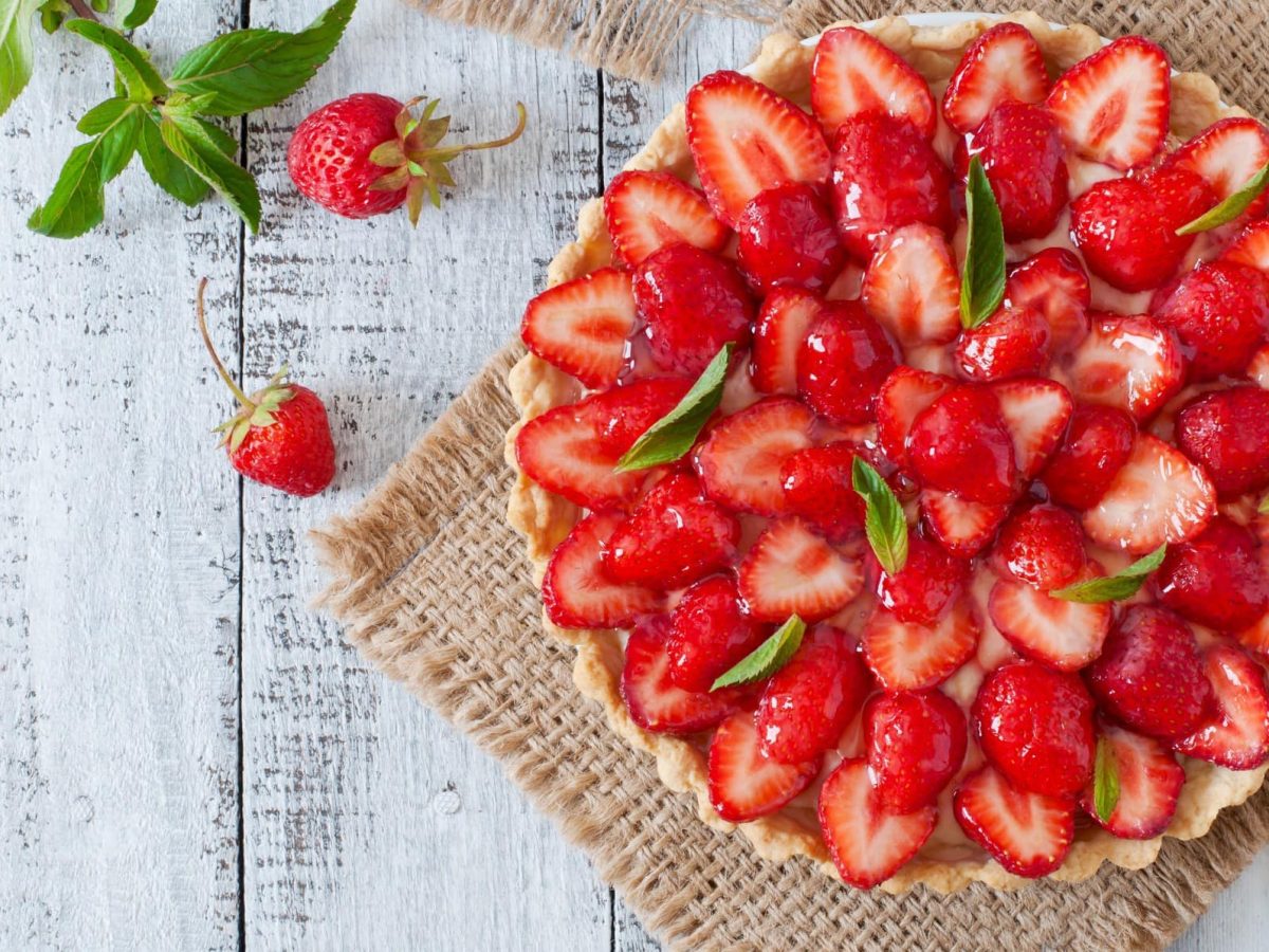 Erdbeerkuchen auf einem geflochtenem Tuch. Daneben Minze und Erdbeeren. Das Ganze auf einem hölzernen Untergrund.