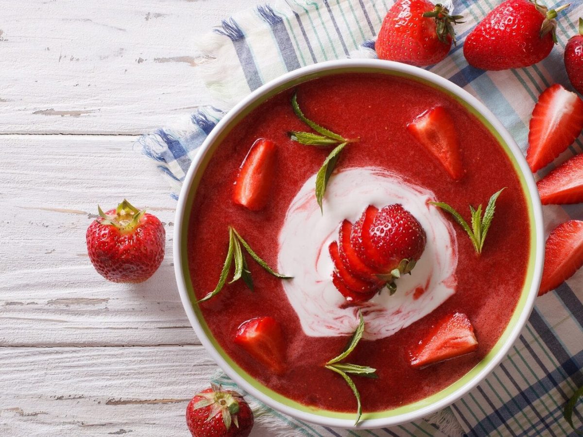 Erdbeersuppe mit frischen Erdbeeren, Quark und Minze in einer weißen Schüssel auf einem weißen Tisch mit einem Geschirrtuch und frischen Erdbeeren.