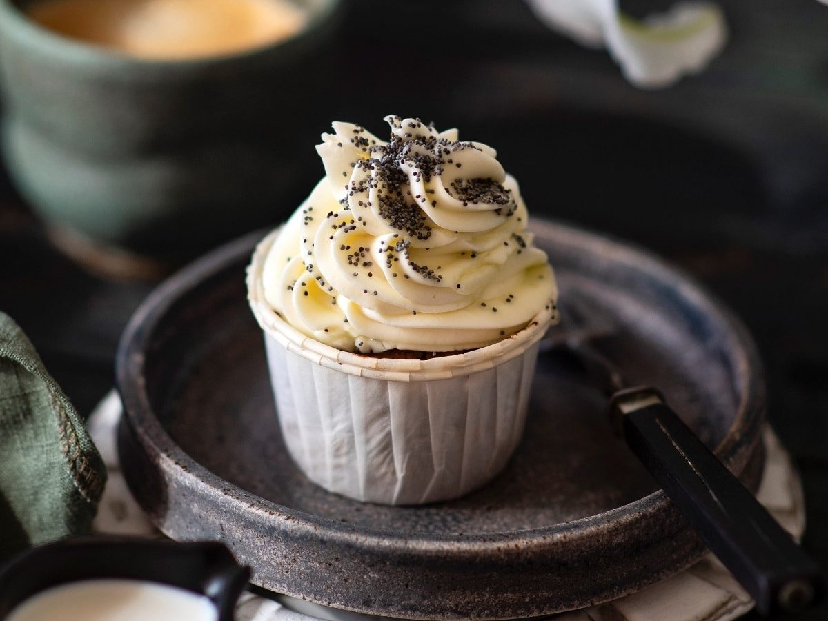 Ein Espresso-Cupcake mit Mohn auf einem dunkelgrauen Teller mit einer Gabel im Vordergrund, sowie eine Tasse Kaffee im Hintergrund.