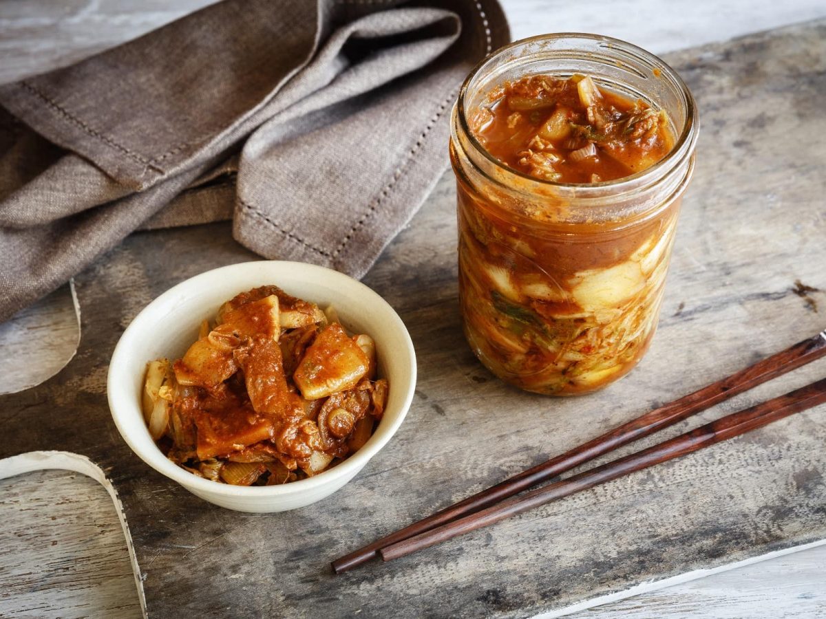 Ein Glas mit fermentiertem Kimchi steht neben einer Schale Reis und Kimchi, daneben liegen Stäbchen.