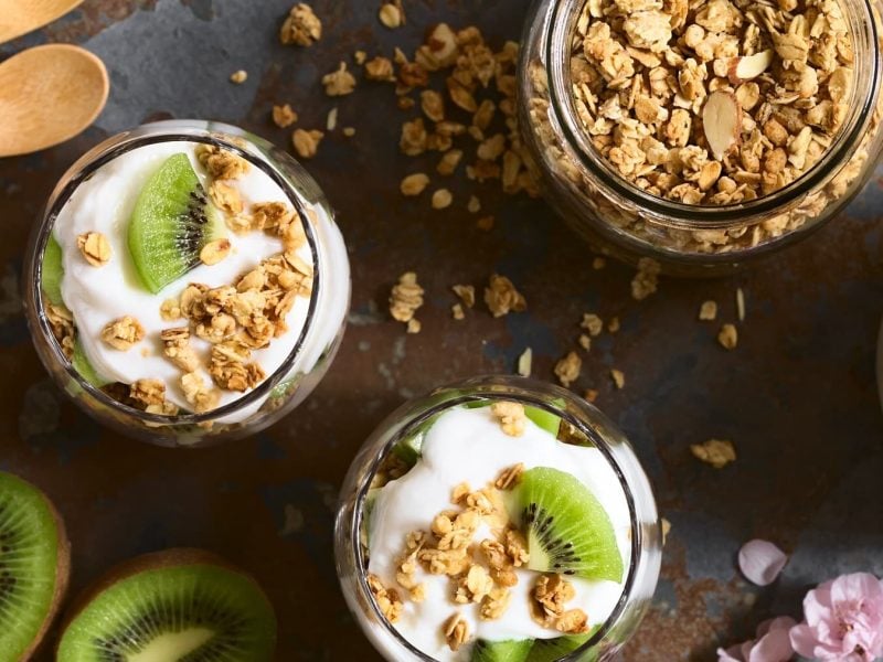 Frühstücks-Skyr mit Granola und Kiwi von Oben in Gläsern neben frischer Kiwi und Blüten auf einem grauen Steintisch