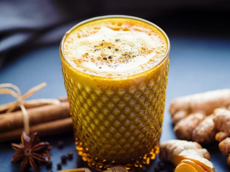 Goldene Milch in einem Glas auf einer Schieferplatte neben Kurkuma, Zimt und Anis vor einem dunklen Tuch.