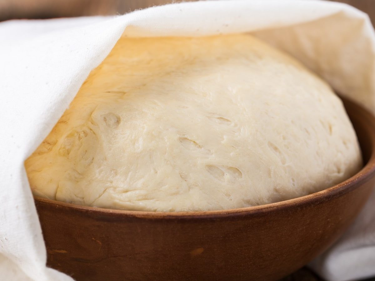 Eine Kugel Hefeteig wird gehen gelassen: Der Teig befindet sich in einer Keramik-Schüssel und ist zur Hälfte mit einem Tuch bedeckt.