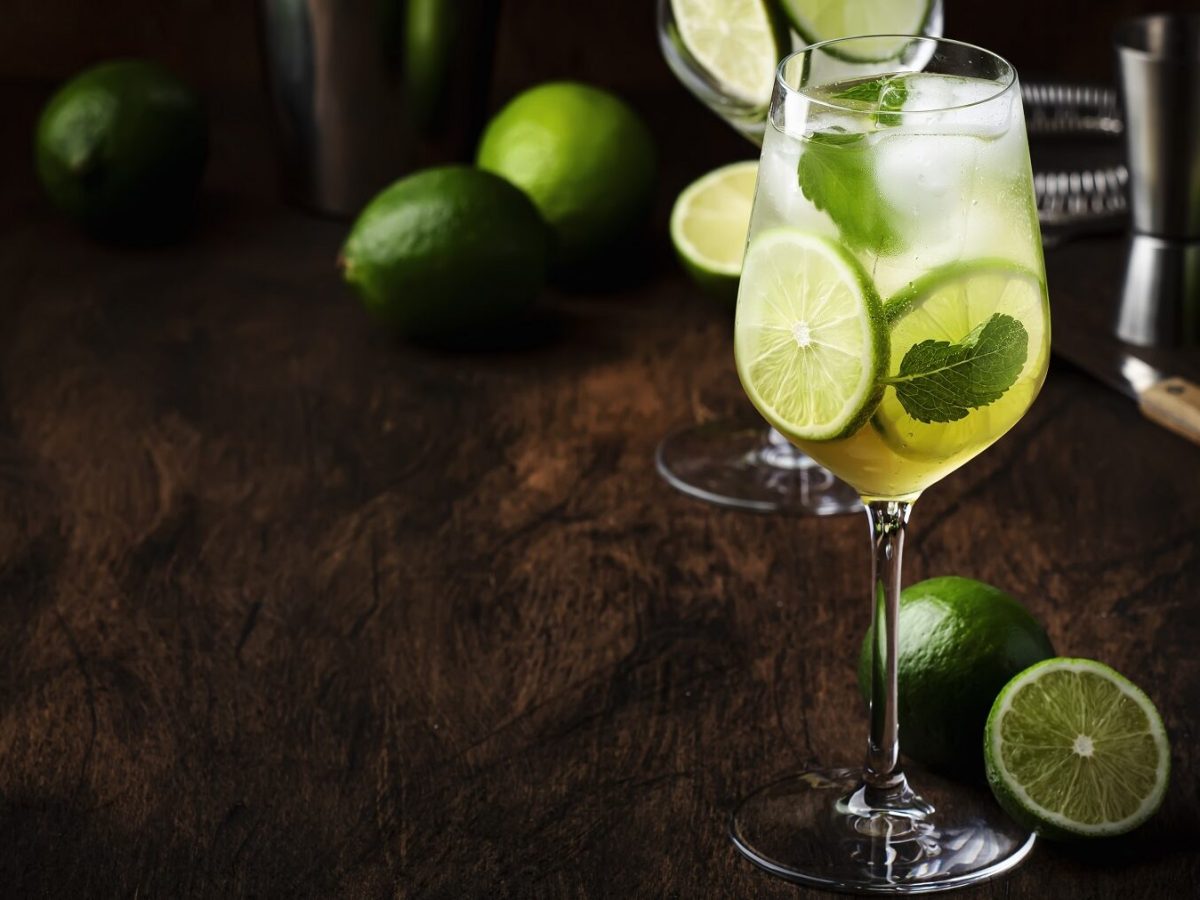 Selbstgemachter Hugo mit Limette und Eis in einem Glas vor einem dunklen Hintergrund mit Cocktail Equipment und frischen Limetten.