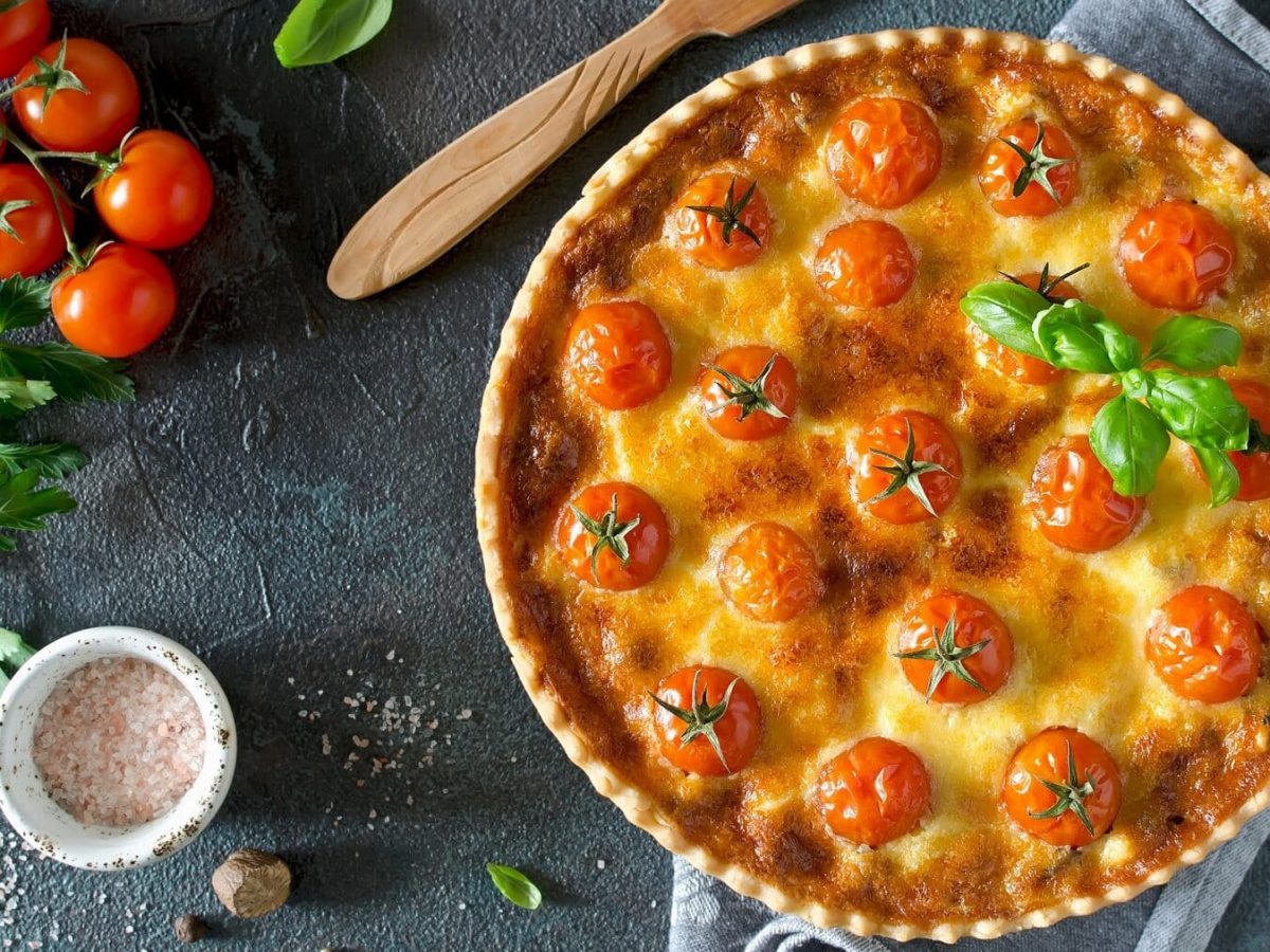 Eine runde Kirschtomaten-Quiche steht auf einem grauen Handtuch auf einem grauen Untergrund. Neben der Quiche liegt ein brauner Holzlöffel. Auf dem Untergrund sind Basilikum, Kirschtomaten und Salzkörner verteilt. Etwas Petersilie liegt neben den Tomaten. Die Quiche ist mit einigen Basilikumblättern angerichtet.