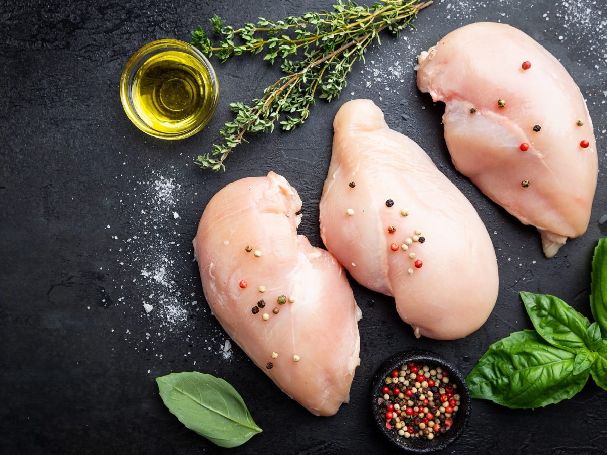 Rohes Hähnchenfleisch für eine Farce von oben auf einem schwarzen Tisch mit frischen Pfefferkörnern, Basilikum, Salz, Thymian und einem Glas Öl.