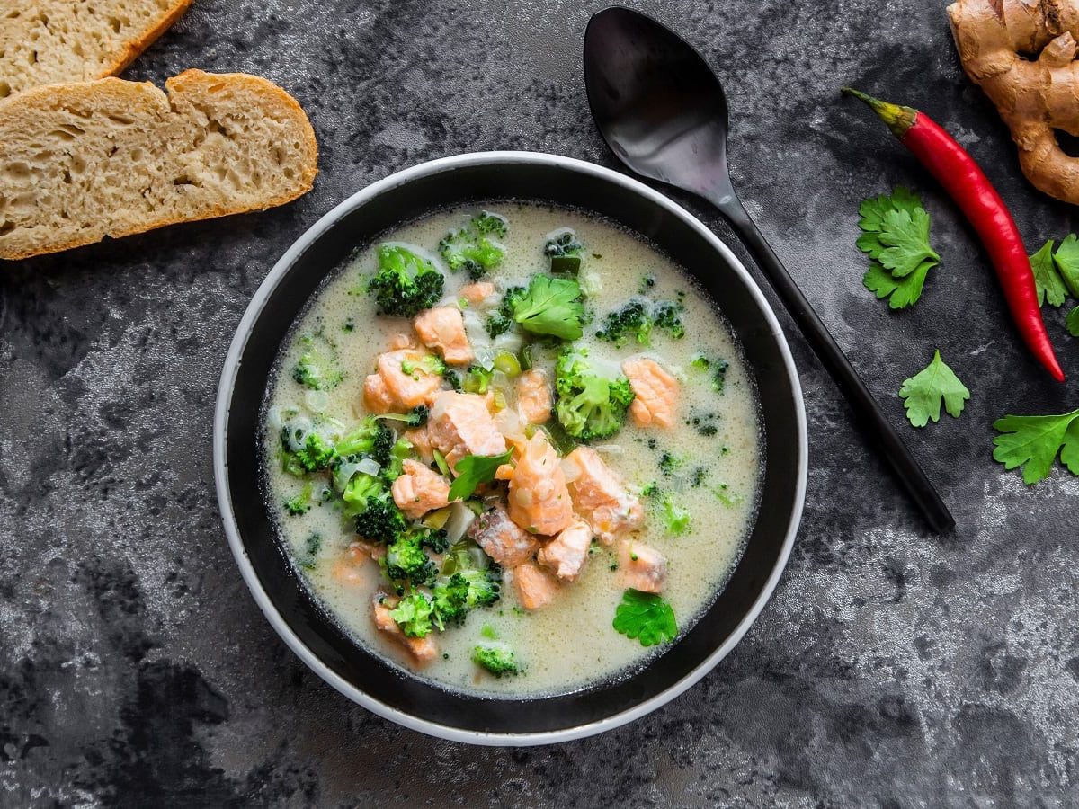 Eine Schüssel mit der Kokos-lachssuppe mit Brokkoli von oben vor grauem Hintergrund neben einer Chilischote, Koriander, Ingwer und zwei Baguette Stücken.