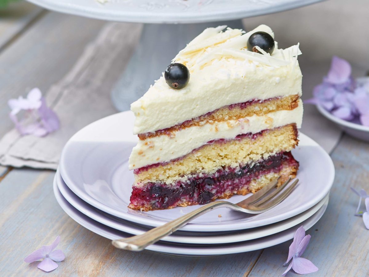 Stück einer Kokostorte mit Johannisbeeren auf weißen Tellern mit einer Gabel auf einem hellen Tisch mit violetten Blüten.