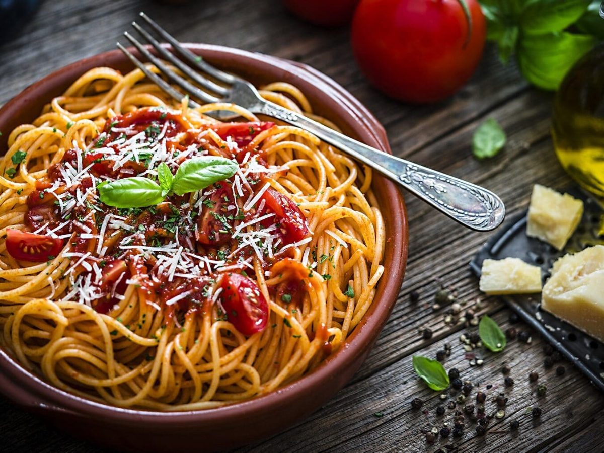 Spaghettisalat mit Tomatensoße, frischem Basilikum und geriebenem Parmesan in einer braunen Tonschüssel auf einem dunklen Holztisch.