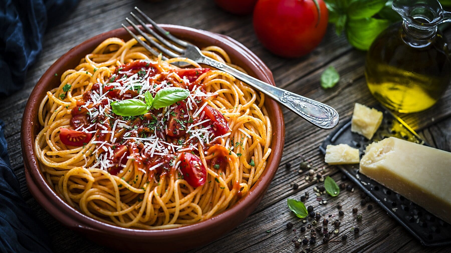 Spaghettisalat mit Tomatensoße, frischem Basilikum und geriebenem Parmesan in einer braunen Tonschüssel auf einem dunklen Holztisch.