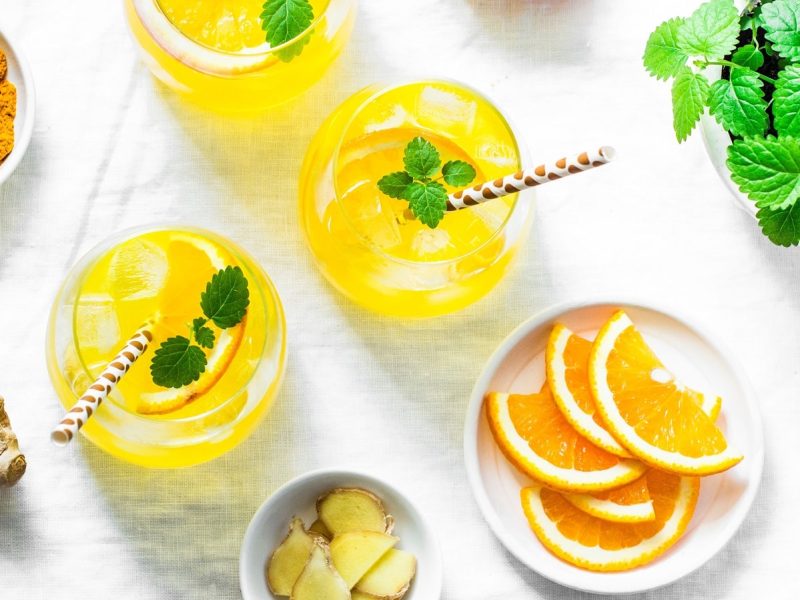 Zwei Gläser gefüllt mit Limonade mit Kurkuma und Inger und garniert mit Orangenscheiben und Minze, neben einer Schale Kurkuma-Pulver, Ingwerscheiben und Orangenscheiben, von oben.