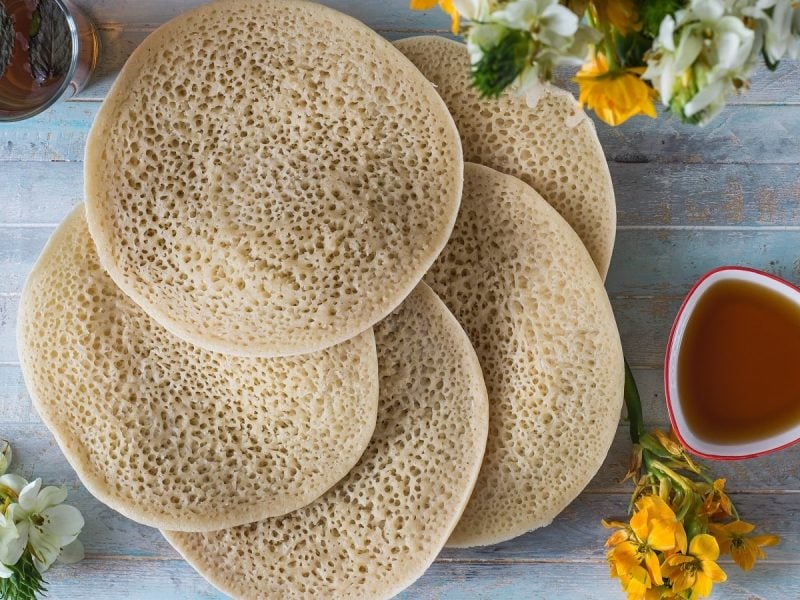 Fünf Baghrir, marokkanische Pfannkuchen aus Grieß mit Luftbläschen auf der Oberseite und fluffigem Teig, liegen gefächert aufeinander, neben einer Schüssel Honigsirup und einem Glas Minz-Tee, von oben vor hellblauem Hintergrund.