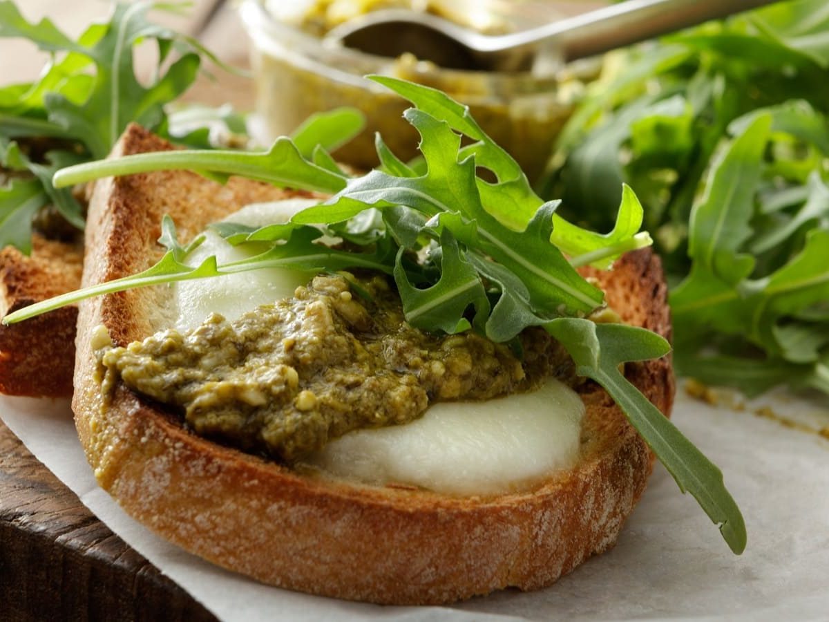 Zwei mit Ziegenkäse überbackene Crostini mit grünem Pesto und Rucola auf einem mit Papier ausgelegten Holztisch von der Seite.