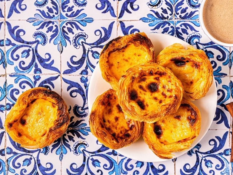 Knusprig gebackene Pastéis de Nata auf einem weißen Teller gestapelt neben einer Tasse Kaffee und zwei Zimtstangen, von oben, vor einem Hintergrund mit portugiesischen Fliesen.