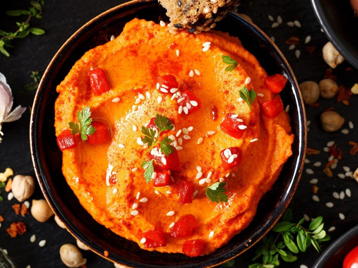 Pikanter Paprika-Hummus mit Sesam in einer dunklen Schale. Daneben einige Gewürze auf einem dunklen Untergrund.