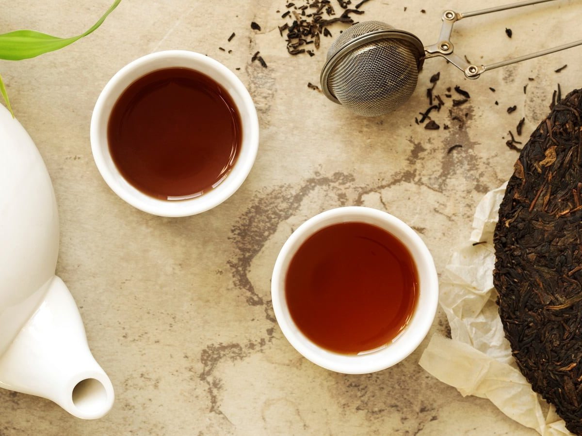 Zwei weiße Tassen mit dunklem Pu Erh-Tee neben einer weißen Teetasse und einem Stück dunklen Pu Erh Tee-Kuchen, vor einem Marmorhintergrund, von oben.