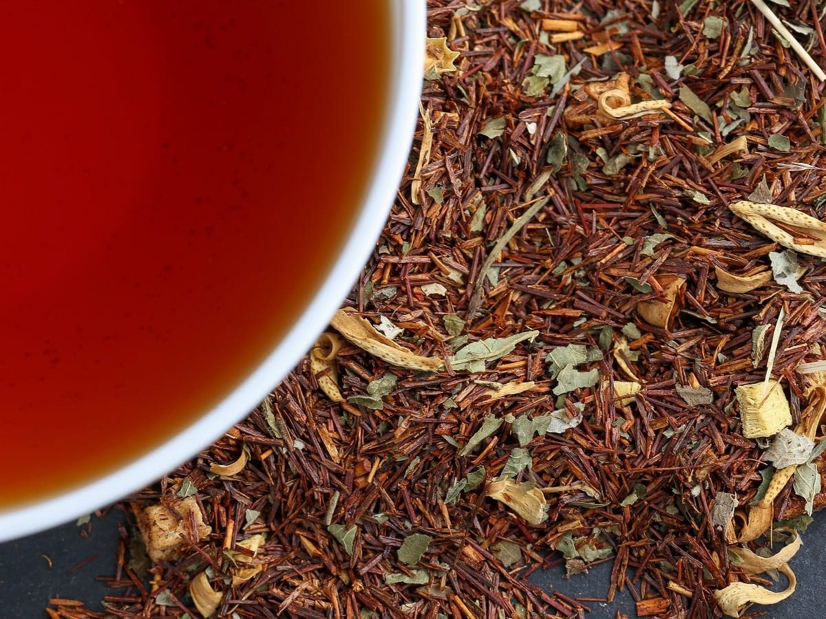 Eine weiße Tasse roter Rooibostee neben getrockneten Teeblättern.