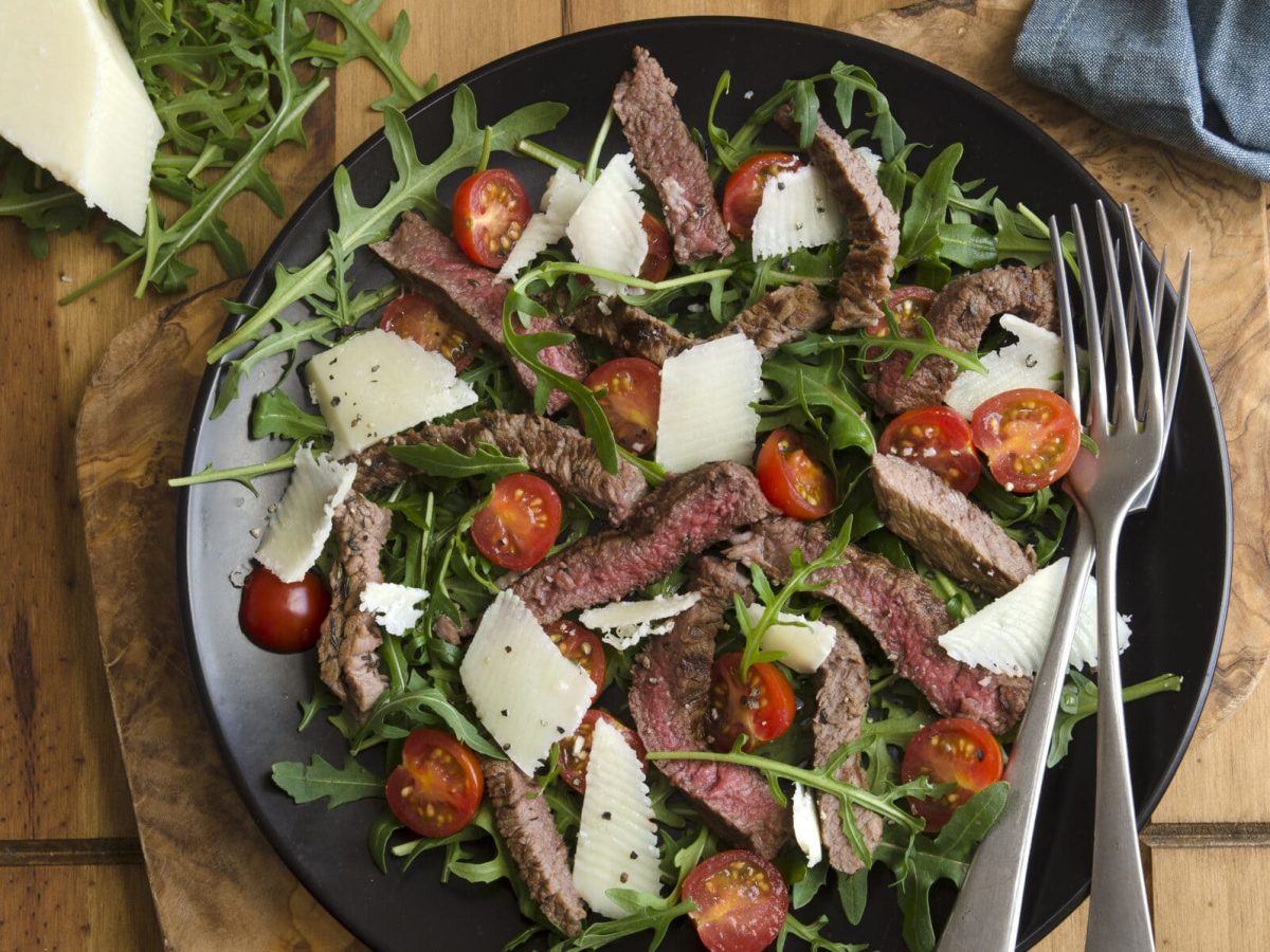 Rumpsteak mit Tomaten-Rucolasalat mit Parmesan in einem tiefen Teller, daneben zwei Gabeln, ein Stück Parmesan und ein Geschirrtuch