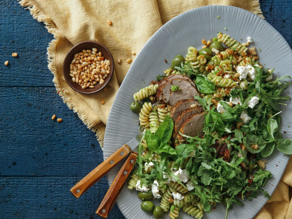 Schweinefilets mit griechischem Nudelsalat und Basilikum auf einem blauen Teller, darunter ein Tuch und daneben eine Schale mit Pinienkernen. Alles auf dunklem Untergrund.